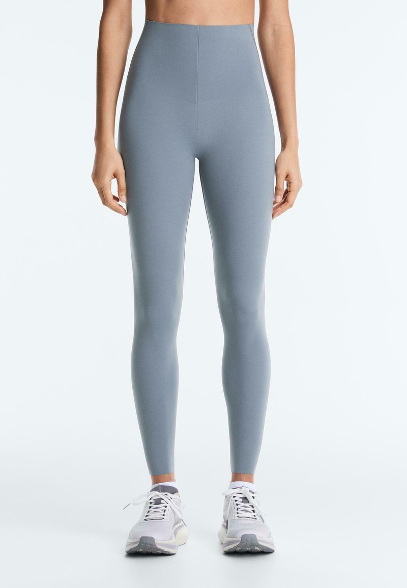 Woman wearing high-waist, light blue leggings and white athletic shoes, standing with arms relaxed at sides on a plain background.