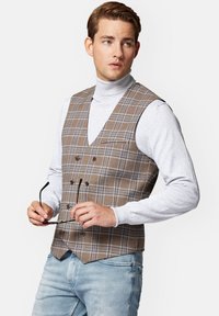 Brązowa kamizelka w kratę z dekoltem w serek, podwójnie zapinana, w połączeniu z jasnoszarym golfem i niebieskimi dżinsami. Okulary trzymane w ręku.