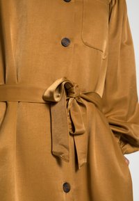 Vestido de camisa em satin castanho com um cinto atado, grandes botões e um único bolso no peito. Apresenta mangas longas e uma textura suave.