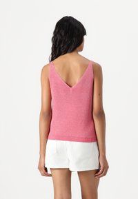 Haut sans manches rose avec un dos en V et un ourlet côtelé. Le matériau a une finition légèrement texturée et brillante. Porté avec un short blanc.