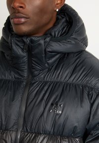Sort pufferjakke med høj krave, hætte og lynlås. Bemærkelsesværdig skinnende adidas-logo på brystet, kviltet tekstur og slankt design.