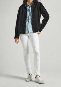 Svart denimjacka med knäppning framtill, kombinerad med en ljusblå chambray-skjorta, vita slim-fit jeans och vita sneakers med grå detaljer.