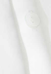 Biała tkanina z widocznym detalem guzika. Tekstura wydaje się gładka z lekkim połyskiem, pokazując czyste, wyprasowane wykończenie i białe przeszycia.