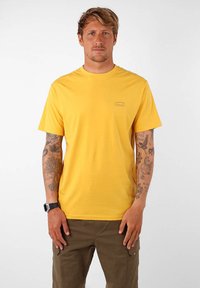 T-shirt en coton jaune avec un col rond et des manches courtes. Présente un petit logo sur la poitrine gauche. Porté avec un pantalon cargo vert.