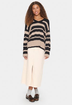 Schwarz-beige gestreifter Pullover mit langen Ärmeln, kombiniert mit einem hellbeigen knöchellangen Rock mit einem Schlitz vorne und braunen Schuhen.