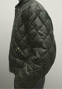 Kurtka bomberpikowana w ciemnozielonym kolorze, z błyszczącym wykończeniem, zippered po przodzie oraz boczn�ą kieszenią z mosiężnym zapięciem na zatrzask.