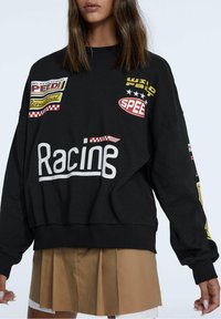 Sudadera negra de gran tamaño que presenta varios parches coloridos con temática de carreras y el texto blanco bordado "Racine" en el frontal.