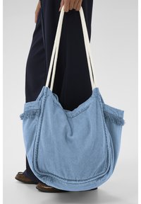 Käsi hoiab suuret, pehmet sinist kangast kandekotti, millel on valged nöörist sangad; allpool on nähtavad tumedad püksid ja pruunid kingad.