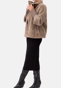 Chaqueta de piel sintética marrón claro con textura, cuello alto, grandes botones y bolsillos laterales, combinada con una falda negra hasta la rodilla y botas negras de tacón.