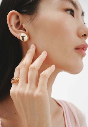 Pendientes de oro en forma de tuerca y un anillo de oro con una piedra preciosa roja, llevados en la mano, contra un fondo neutro. Textura suave y formas redondeadas.