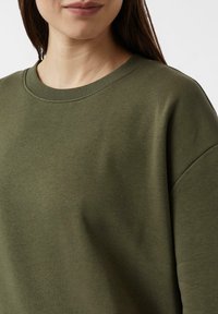 Bluza w kolorze oliwkowej zieleni z okrągłym dekoltem i żebrowanymi detalami. Miękka, teksturowana tkanina o luźnym kroju i lekko opuszczonych ramionach.