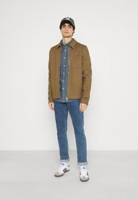 Bruin wollen mixjacket met een grijs shirt, blauwe jeans en witte sneakers. Bevat een borstzak en opgerolde mouwen. Draagt een groene pet.