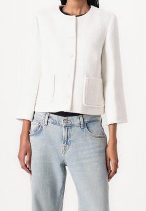 Femme portant une veste blanche texturée boutonnée avec deux poches avant et un jean taille haute bleu clair sur fond blanc.