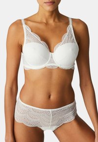 Vit spets-bh med djup urringning och scalloperade kanter, i kombination med matchande spets-trosa som har ett vågigt mönster vid fållen.