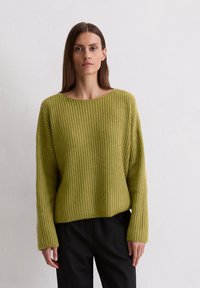 Groene gebreide trui met een losse pasvorm, wijde halslijn en ribbelstructuur. Gepaard met zwarte broek, voorzien van lange mouwen.