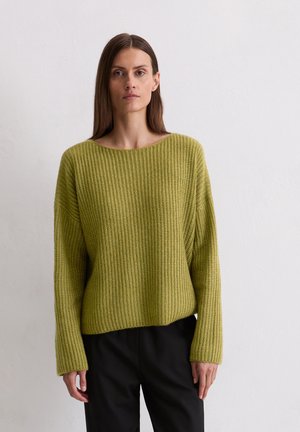 Groene gebreide trui met een losse pasvorm, wijde halslijn en ribbelstructuur. Gepaard met zwarte broek, voorzien van lange mouwen.