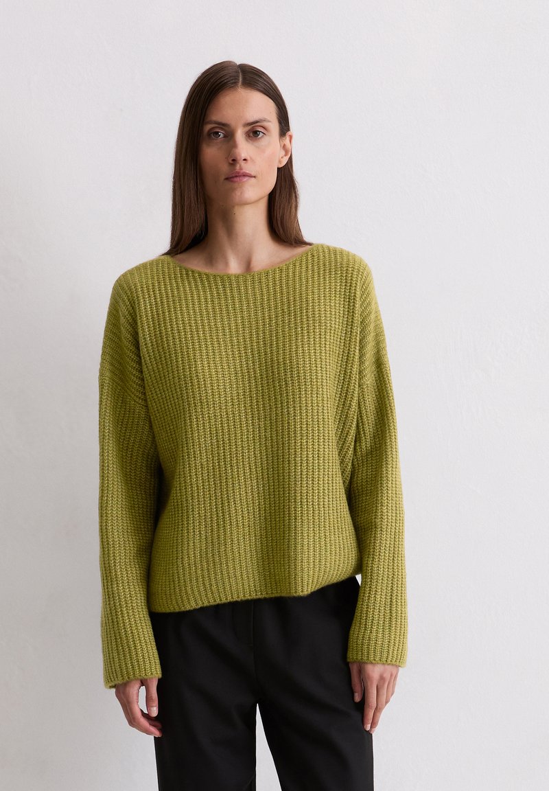 Groene gebreide trui met een losse pasvorm, wijde halslijn en ribbelstructuur. Gepaard met zwarte broek, voorzien van lange mouwen.