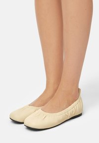 Beige soft leather ballet flats with gathered elastic edging worn on bare feet against a white background.