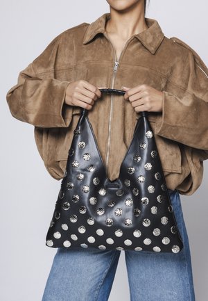 Osoba v hnedom semišovom saku a modrých džínsoch drží čiernu koženú kabelku so striebornými kruhovými cvočkmi za zabalenú rukoväť.