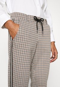Pantalon à motif pied-de-poule marron et beige avec une taille à cordon, des poches latérales et des bandes latérales noires contrastantes.