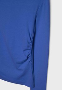 Modri dolg rokav z detajlom nabiranja na strani in gladko teksturo tkanine na enostavni svetli podlagi.