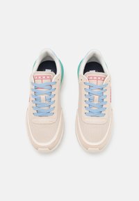 Zapatillas con una parte superior de lona beige, cordones azules y acentos verdes en la suela. Presenta un parche de logo rojo en la lengüeta y un diseño texturizado.