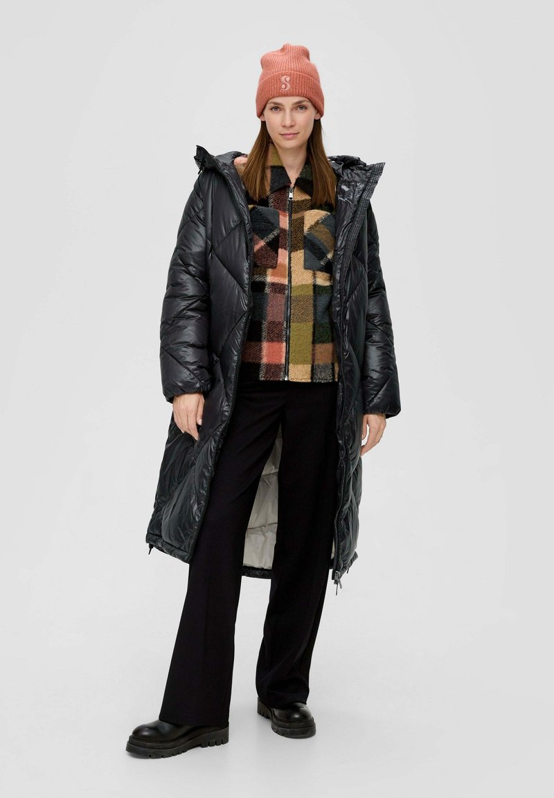 Langer schwarzer Daunenmantel mit einem Rautenmuster, getragen über einer karierten Fleecejacke. Schwarze Hose und klobige schwarze Stiefel runden das Outfit ab.