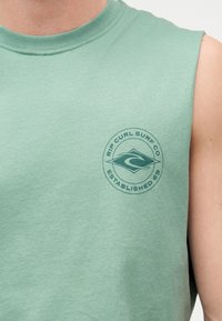 Πράσινο αμάνικο πουκάμισο που φοριέται από ένα άτομο, με κυκλικό λογότυπο "Rip Curl Surf Co Established 69" στο στήθος.