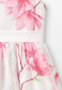 Květinové šaty z bílé látky s růžovými motivy magnólií a puntíky, doplněné bílým pasem a nařasenou sukní.