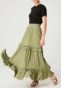 Jupe maxi en vert olive à volants, en tissu texturé léger, associée à un t-shirt noir ajusté et des sandales noires à lanières.