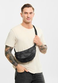 Zwarte camouflage schoudertas met een zachte textuur, verstelbare band en logo-detail. Draagt over het lichaam door een persoon in een lichte blouse.