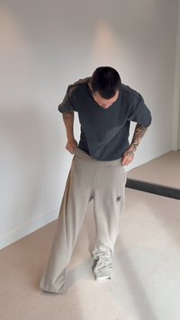 Mann med tatoveringer som tar på seg store beige bukser i et minimalistisk rom med hvite vegger og lyst gulv.