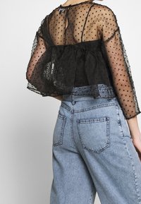 Top in rete nera con pois, design peplo, maniche lunghe trasparenti e schiena scolpita. Indossato con jeans a vita alta di colore blu chiaro.