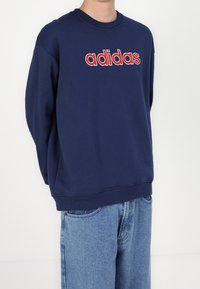 Person som bär en marinblå Adidas-tröja med röd och vit logotyp, tillsammans med ljusblå jeans mot en enfärgad vit bakgrund.