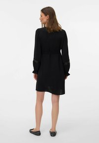 Schwarzes Langarmkleid mit taillierten Bund, rüschierten Bündchen und Spitzenakzenten. Weicher Stoff und knielanger Schnitt.