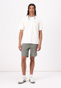 T-shirt blanc à manches courtes avec logo sur la poitrine, associé à un short olive vert texturé et des baskets blanches et noires.