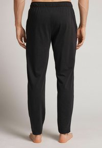 Zwarte loszittende broek met een elastische tailleband, gladde textuur, taps toelopende pijpen en geen zichtbare zakken of patronen. Blootsvoets zichtbaar.
