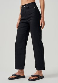 Visoko pasu črne kavbojke s ravno nogo, ki vključujejo sprednje žepi in srebrne gumbne dodatke. Dopolnjene s črnimi sandali.