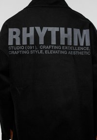 Zwarte stoffen shirt met grote grijze tekst op de achterkant die leest: "RHYTHM STUDIO (091), UITMUNTENDHEID VERVAARDIGEN, STIJL VERVAARDIGEN, ESTHETIEK VERHOGEN."