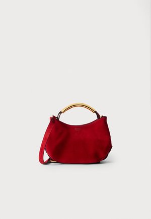 Sac à main en daim rouge avec une forme courbée, anse en métal doré et bandoulière amovible. Texture lisse, design minimaliste et détail du logo.
