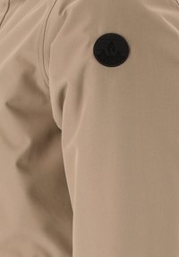 Ljust beige jacktyg med en slät yta och matt finish, med en rund svart logotyp-patch på överarmen.