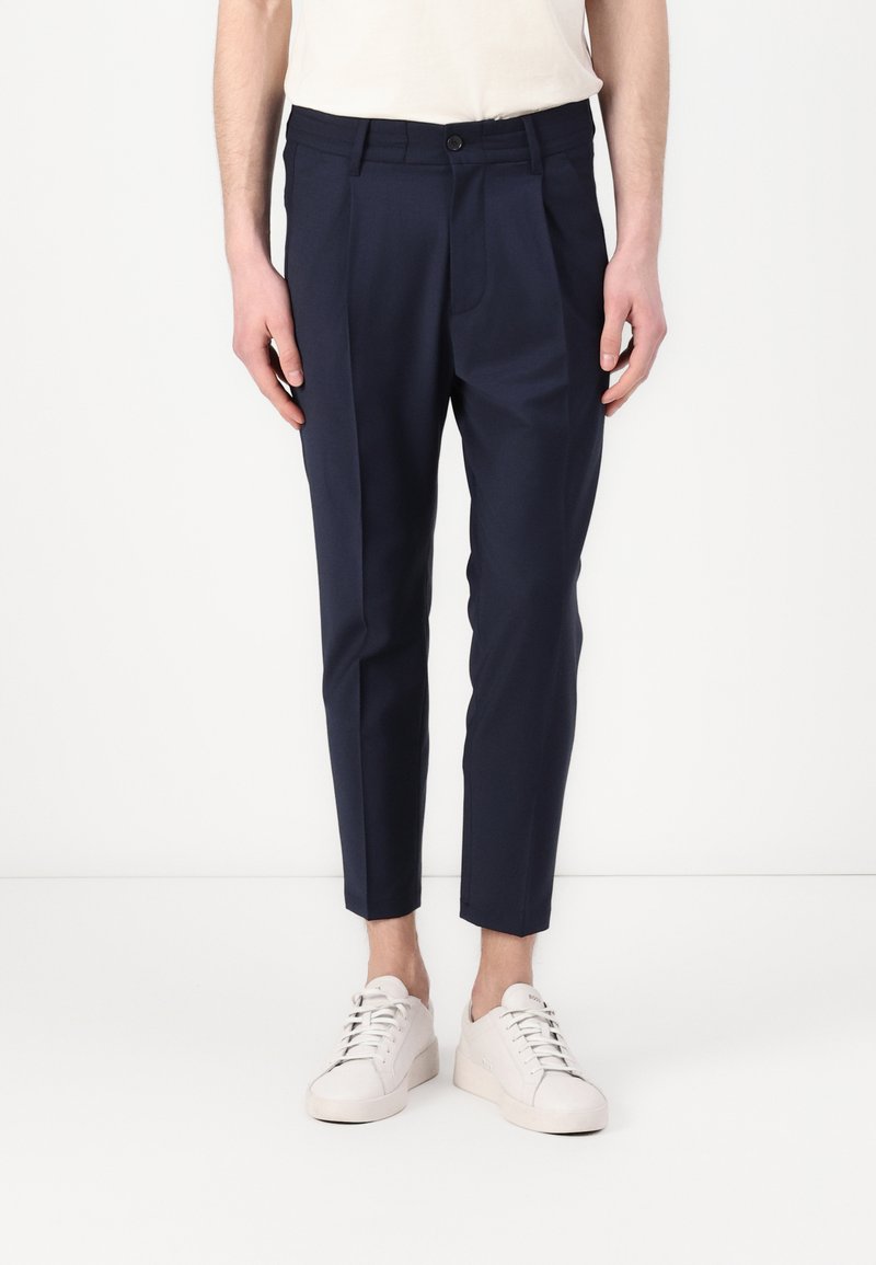 Pantalon marine à coupe ajustée, doté de plis sur le devant, de poches latérales et d'une texture lisse. Porté avec des baskets blanches.