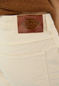 Šviesiai rudos medvilnės kelnės su rudos odos Dickies logotipo užpakaliu, matomu siuvinėjimu ir kišenėmis.