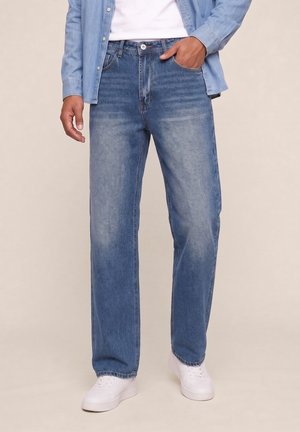 Homme portant un jean bleu délavé droit, des baskets blanches, une chemise blanche et une veste en denim bleu clair, main dans la poche.