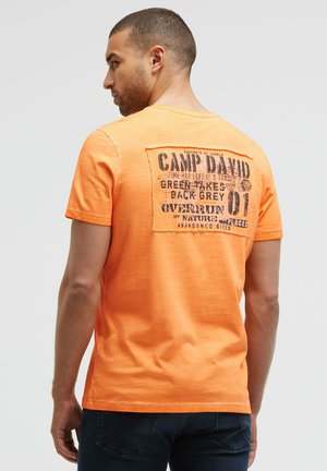 Mann, der mit dem Rücken zur Kamera steht, trägt ein oranges T-Shirt mit kurzen Ärmeln und einem schwarzen bedruckten Schriftzug auf dem oberen Rücken sowie dunkle Jeans.