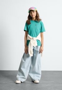 Mėlynas marškinėliai su kišenėle krūtinėje, kreminis megztinis surištas aplink liemenį, šviesiai mėlyni plačios kojų džinsai ir daugiaspalvė kepurė.