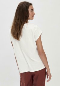 Weißes T-Shirt mit kurzen Ärmeln, entspannter Passform und umgeschlagenen Ärmeln, kombiniert mit rustfarbenen Hosen, getragen von einer Person, die von hinten gesehen wird.