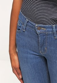 Detailní záběr na osobu v modrých džínách a černobíle pruhovaném topu, s levou rukou položenou na boku.