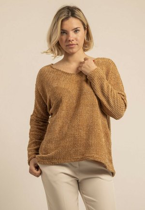 Femme blonde portant un pull en maille marron ample et un pantalon beige, debout devant un fond beige uni.