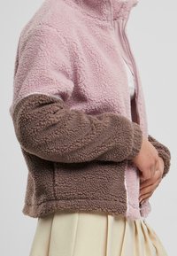Chaqueta de forro polar con paneles rosa y marrón, cierre con cremallera en la parte delantera y tela suave y texturizada. Cuenta con puños elásticos y un diseño corto.